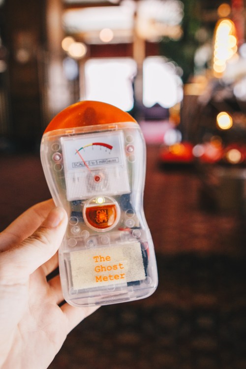 A person holds a transparent device labeled "The Ghost Meter" with a gauge, set against a blurred background.