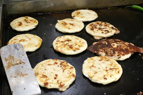 A griddle with several golden-brown pupusas cooking, some with crispy edges, alongside a spatula.