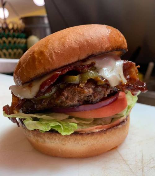 A delicious burger with bacon, cheese, lettuce, tomato, and pickles on a toasted bun, placed on a cutting board.