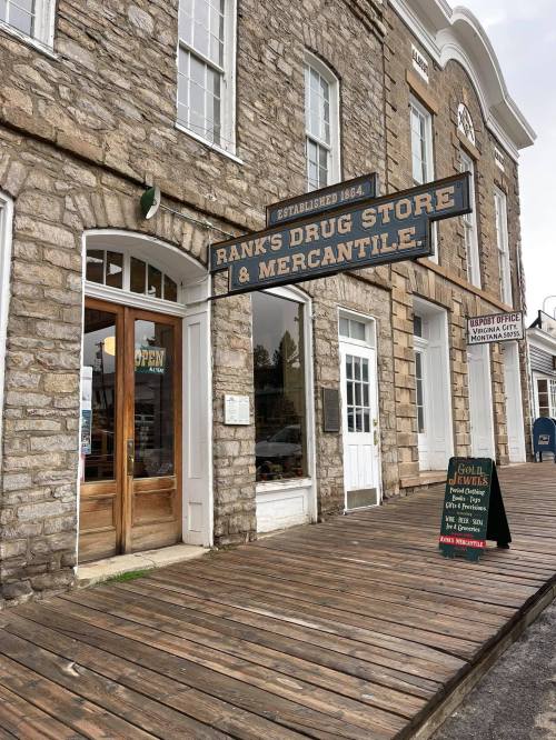 Exterior of Rank's Drug Store & Mercantile, featuring a wooden porch and a sign indicating it's open. Stone building facade.
