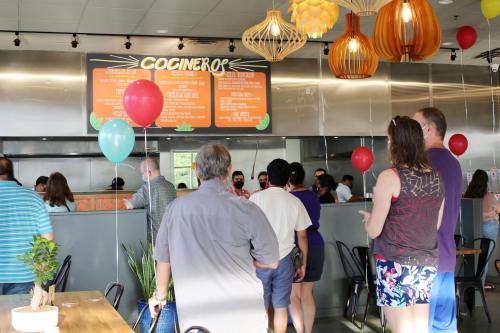 A busy restaurant interior with customers waiting in line, colorful balloons, and decorative lighting overhead.