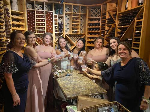 A group of eight women in formal dresses toast with glasses in a wine cellar setting.