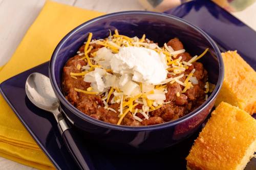 A bowl of chili topped with cheese and onions, served with cornbread on a blue plate and a spoon beside it.