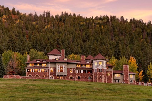 A large, elegant stone mansion surrounded by lush green trees and a colorful sunset sky.