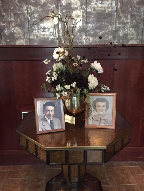 A wooden table displays two framed vintage photos and a floral arrangement against a textured wall.