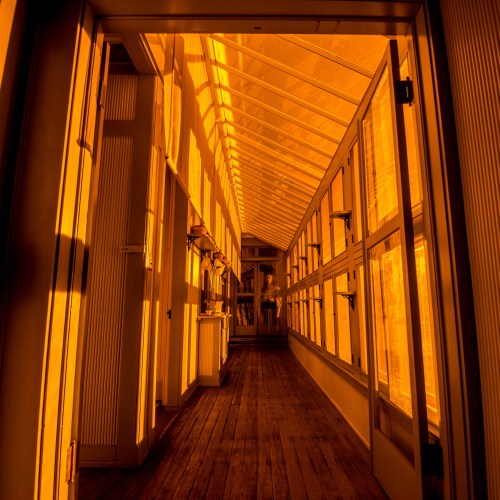 A sunlit corridor with wooden floors and large windows, casting warm orange shadows.