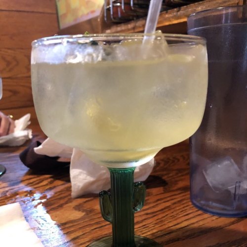 A close-up of a green cocktail in a cactus-shaped glass, with ice and a straw, on a wooden table.