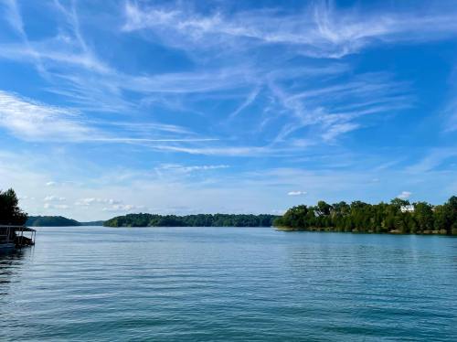 A serene lake scene with calm waters, lush green trees, and a clear blue sky with wispy clouds.