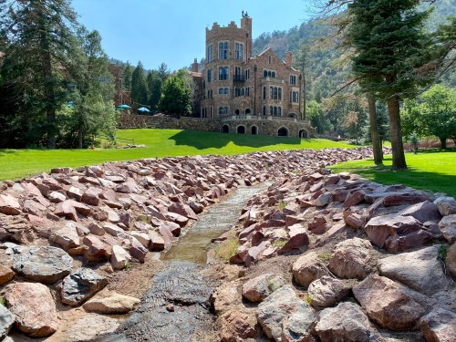 A stone-lined creek flows through a green landscape, with a large, historic building in the background.