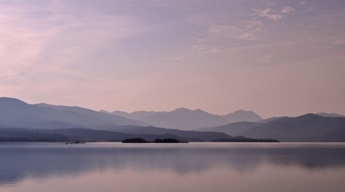 Serene landscape featuring calm water, distant mountains, and a soft pastel sky at dawn or dusk.