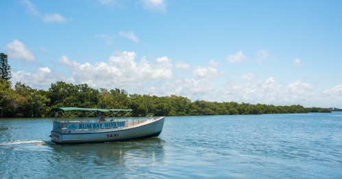 A water taxi labeled "Rum Bay Water Taxi" glides through a calm river surrounded by lush greenery under a blue sky.