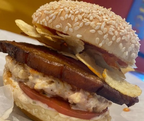 A close-up of a sesame seed bun sandwich with layers of meat, tomato, sauce, and potato chips on top.