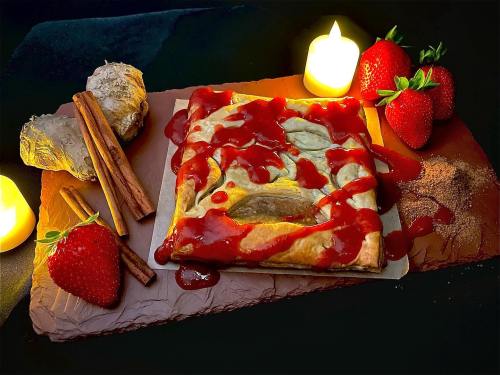 A pastry topped with red sauce, surrounded by fresh strawberries, cinnamon sticks, and candles on a dark background.