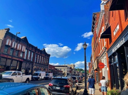A bustling street scene with brick buildings, parked cars, and a clear blue sky with fluffy clouds.