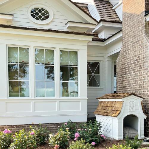 A charming house exterior featuring large windows, a small doghouse, and blooming flowers in the garden.