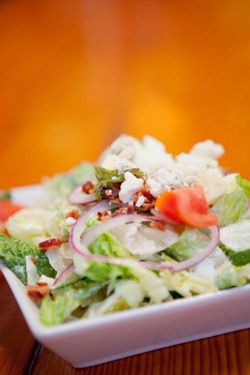 A fresh salad with romaine lettuce, tomatoes, red onions, bacon bits, and feta cheese in a white bowl.
