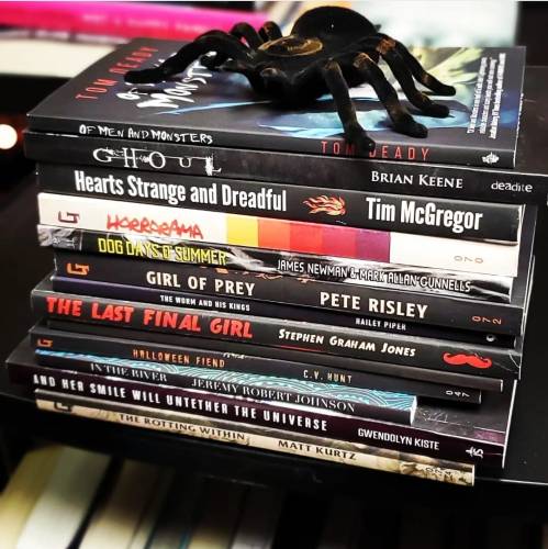 A stack of horror-themed books with a decorative spider on top. Titles include "Ghoul" and "The Last Girl of Grey."