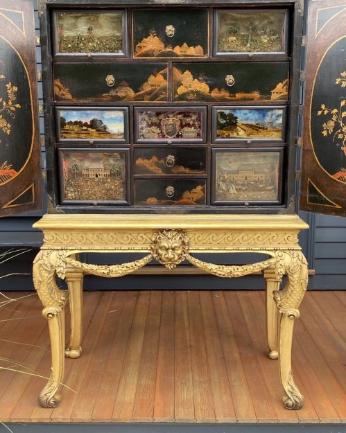 A decorative cabinet with multiple small drawers, featuring intricate landscapes, sits atop an ornate table with lion motifs.