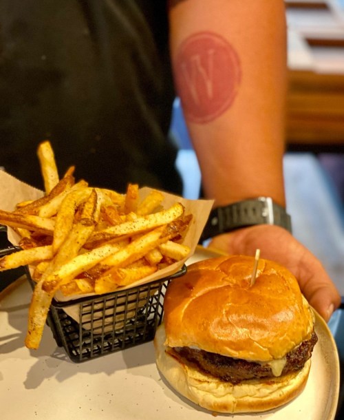 A close-up of a burger with a side of fries, held by a person with a tattoo on their arm.