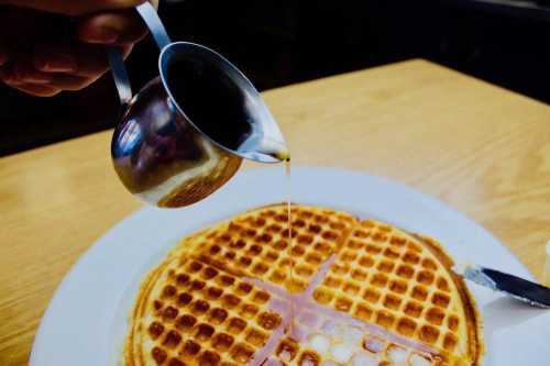 A hand pours syrup from a small metal pitcher onto a golden-brown waffle on a white plate.