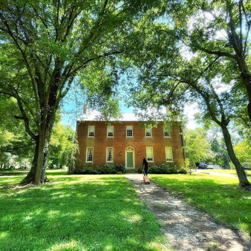 A brick house surrounded by lush green trees and grass, with a person walking along a path towards it.