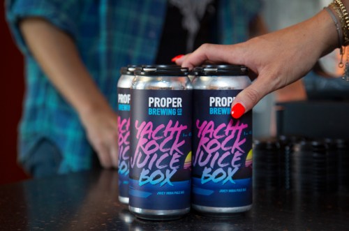A hand reaches for a four-pack of "Yacht Rock Juice Box" beer cans on a bar counter.