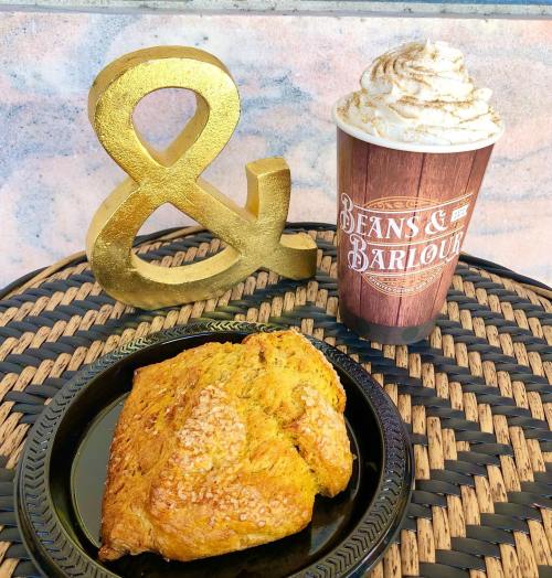 A golden ampersand next to a coffee cup and a scone on a black plate, set on a woven surface.