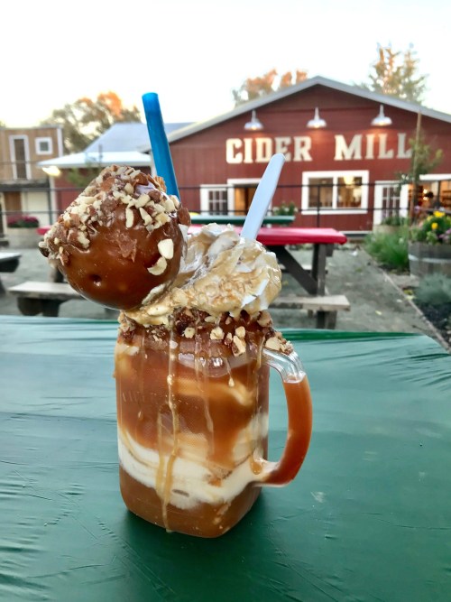 A caramel apple milkshake topped with a caramel apple and nuts, set against a cider mill backdrop.