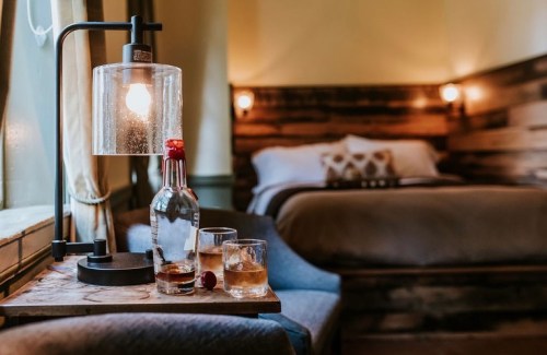 A cozy bedroom with a wooden bed, a lamp, and a table featuring two glasses of whiskey and a bottle.