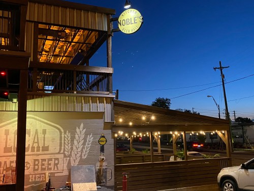 Exterior of Noble's Beer Hall at dusk, featuring a sign, outdoor seating, and warm lights against a blue sky.