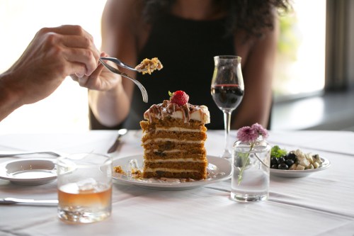 A person enjoying a slice of layered cake with a strawberry on top, accompanied by drinks and a decorative table setting.
