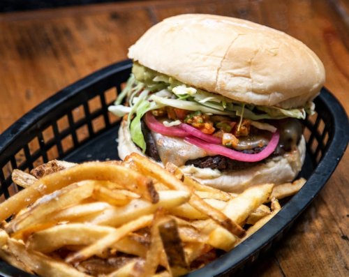 A juicy burger with toppings and pickled onions, served with a side of crispy French fries in a black basket.