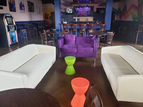 A vibrant lounge area with two white sofas, a purple chair, and a green side table, set in a colorful bar environment.
