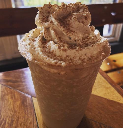 A creamy coffee drink topped with whipped cream and a sprinkle of cocoa, served in a clear cup on a wooden table.