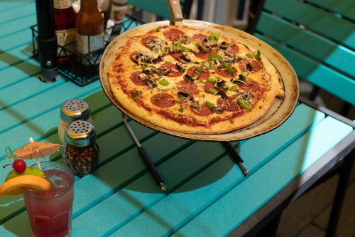A freshly baked pepperoni pizza on a stand, with a colorful cocktail and condiments on a turquoise table.