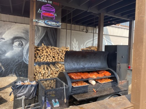 A barbecue smoker filled with ribs and meats, surrounded by stacked firewood and a mural in the background.