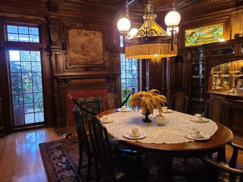 A vintage dining room with a wooden table set for tea, ornate decor, and large windows letting in natural light.