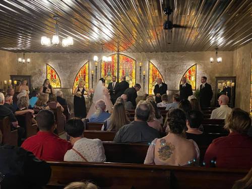 A wedding ceremony in a chapel with stained glass windows, featuring a bride and groom surrounded by guests.