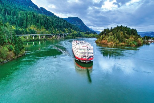A riverboat navigates a serene river surrounded by lush green hills and a cloudy sky.