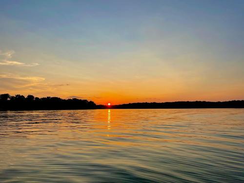 A serene sunset over calm water, with silhouettes of trees along the horizon and vibrant orange and blue hues in the sky.