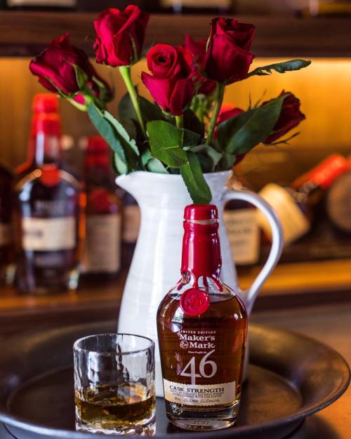 A bottle of Maker's Mark 46 whiskey with a glass, accompanied by a vase of red roses on a tray.