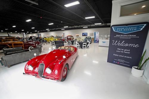 A red classic car in a spacious museum with tables and a welcome banner for events.