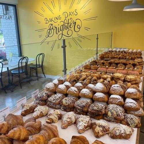 A display of various baked pastries in a bright café, with a wall sign that reads "Baking Days Brighter."