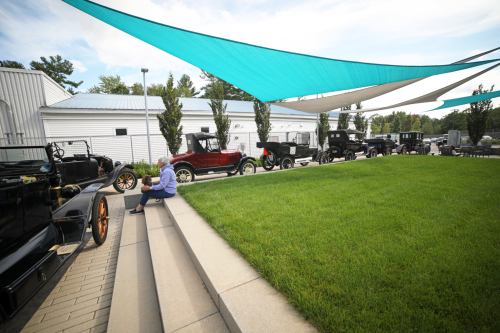 A person sits on steps near vintage cars under colorful canopies, with green grass and a building in the background.