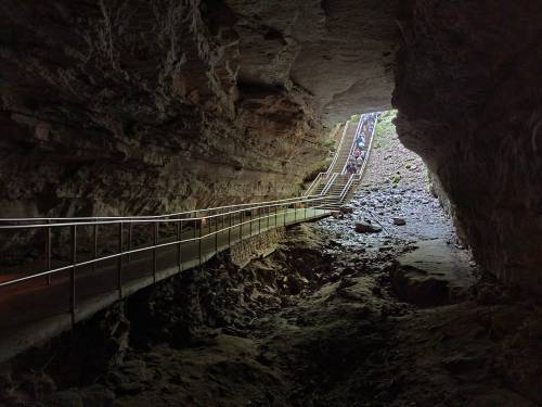 A rocky cave entrance with a metal walkway leading out to bright light and a group of people in the distance.