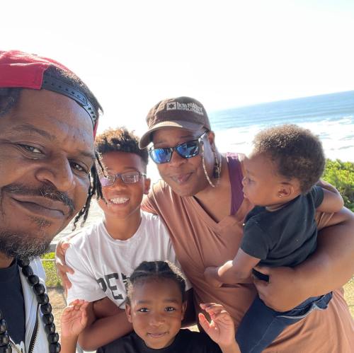 A family poses for a selfie by the ocean, smiling with two children and a baby in their arms.