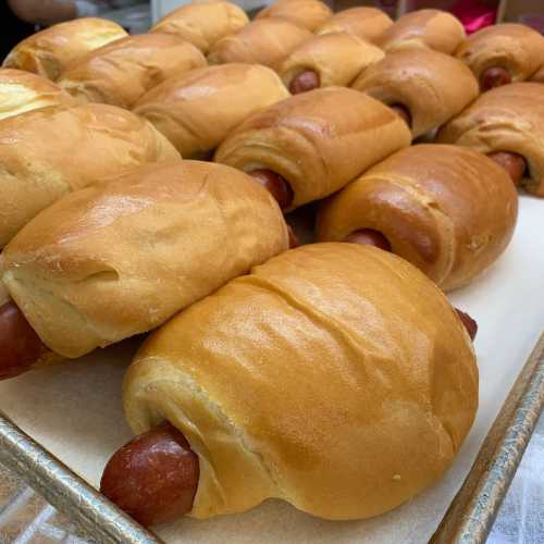 A tray of freshly baked hot dog buns, each with a sausage partially visible inside.