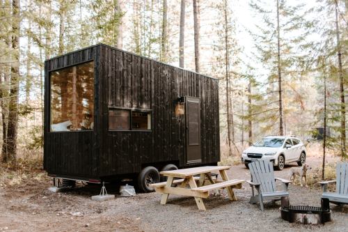 A modern black tiny house on wheels surrounded by trees, with a picnic table and a parked car nearby.