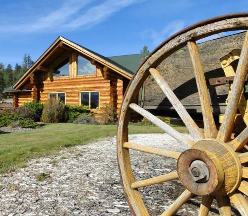 A rustic log cabin with a green roof, framed by a wooden wagon wheel in the foreground and lush landscaping.