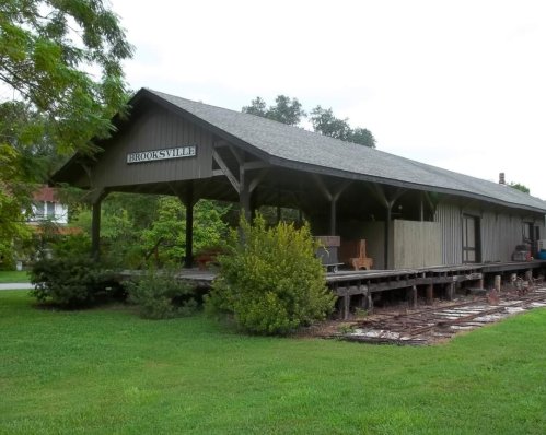 A wooden train station building with a sign reading "BROOKSVILLE," surrounded by greenery and a grassy area.